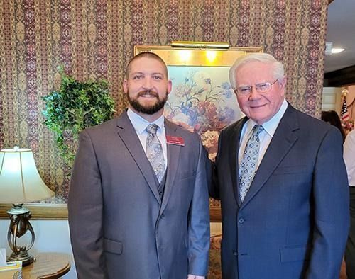 Two men in suits pose, smiling, in front of a floral wallpaper and painting.