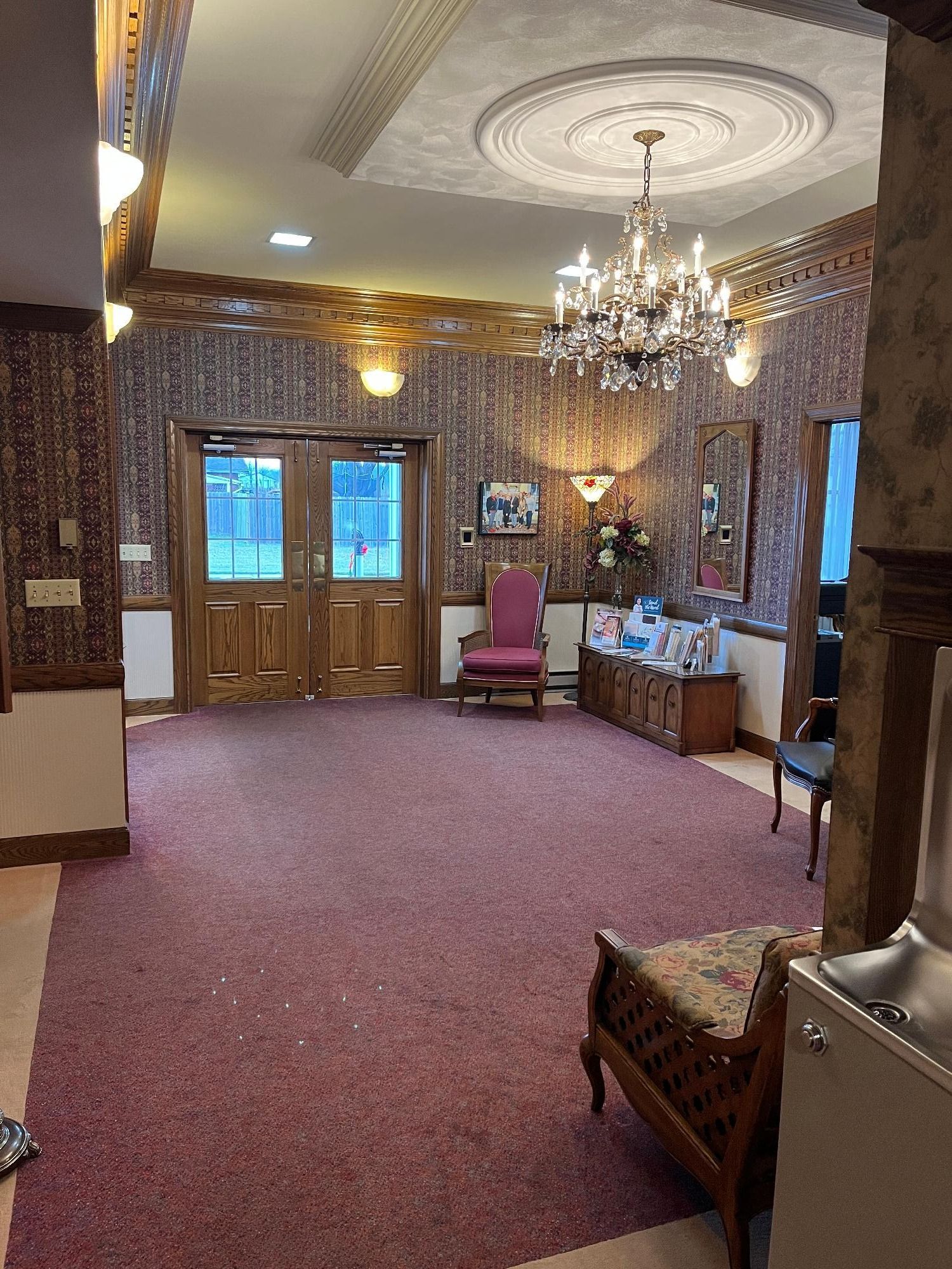 An ornate room with patterned wallpaper, chandelier, red carpet, and double doors.