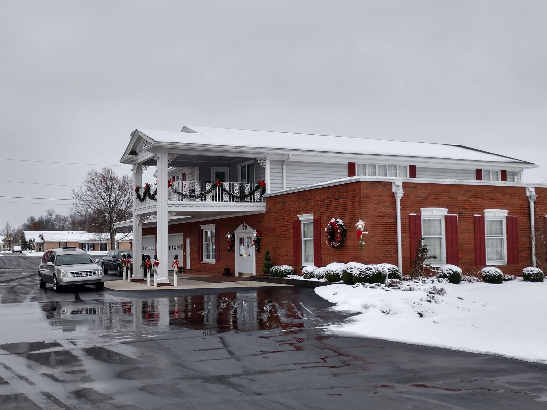Snow-covered building with red brick and white siding; Christmas wreaths visible. Cars parked in front, overcast sky.