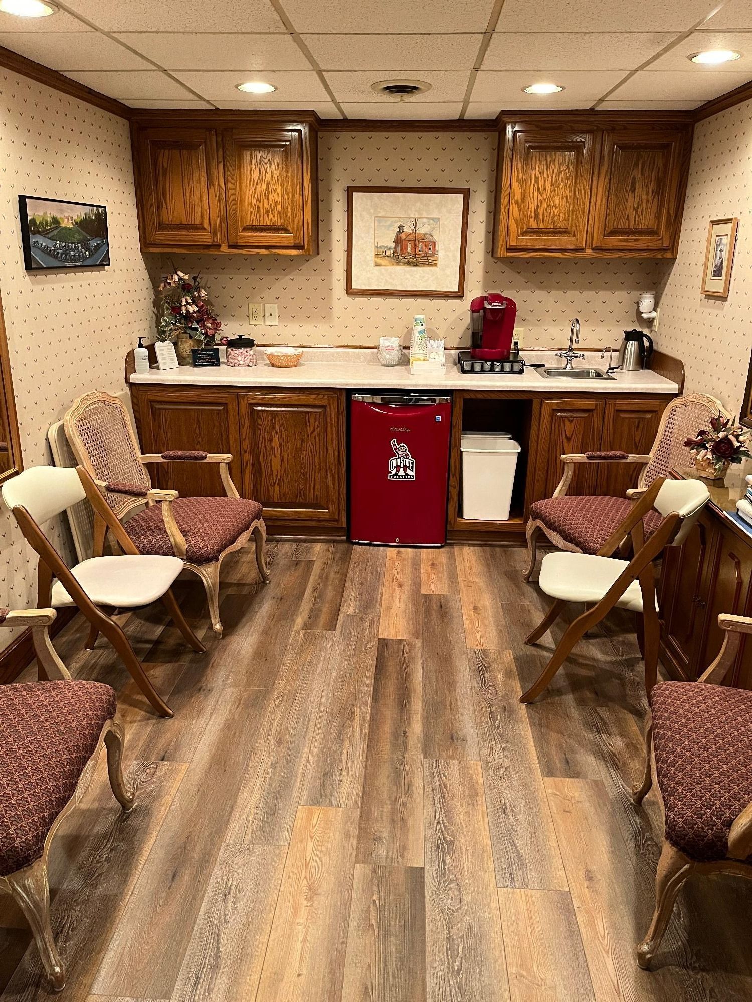 A waiting area with chairs, a counter with coffee maker, and a red mini-fridge. Wooden cabinets.