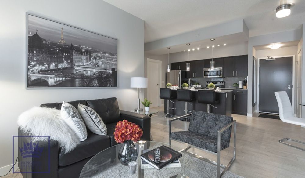 Modern living room with black leather couch, open kitchen with bar seating, cityscape art.