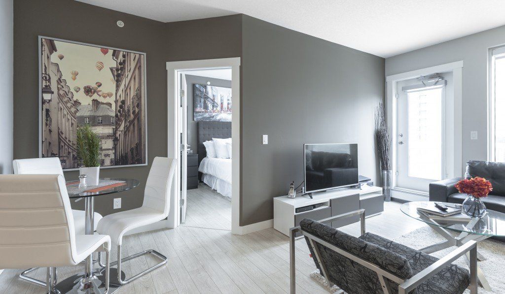 Modern apartment interior with dining area, living room, and bedroom visible. Gray walls, white furniture.