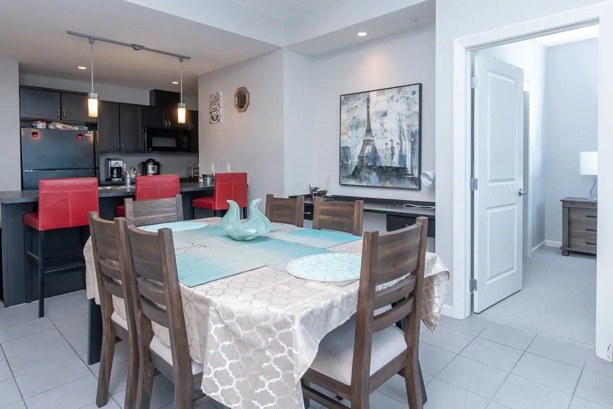 Dining room with table set for six, kitchen in background, artwork on the wall.