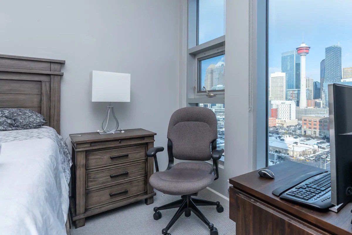 Bedroom with bed, desk, office chair, and city view through large window. Calgary furnished apartments 