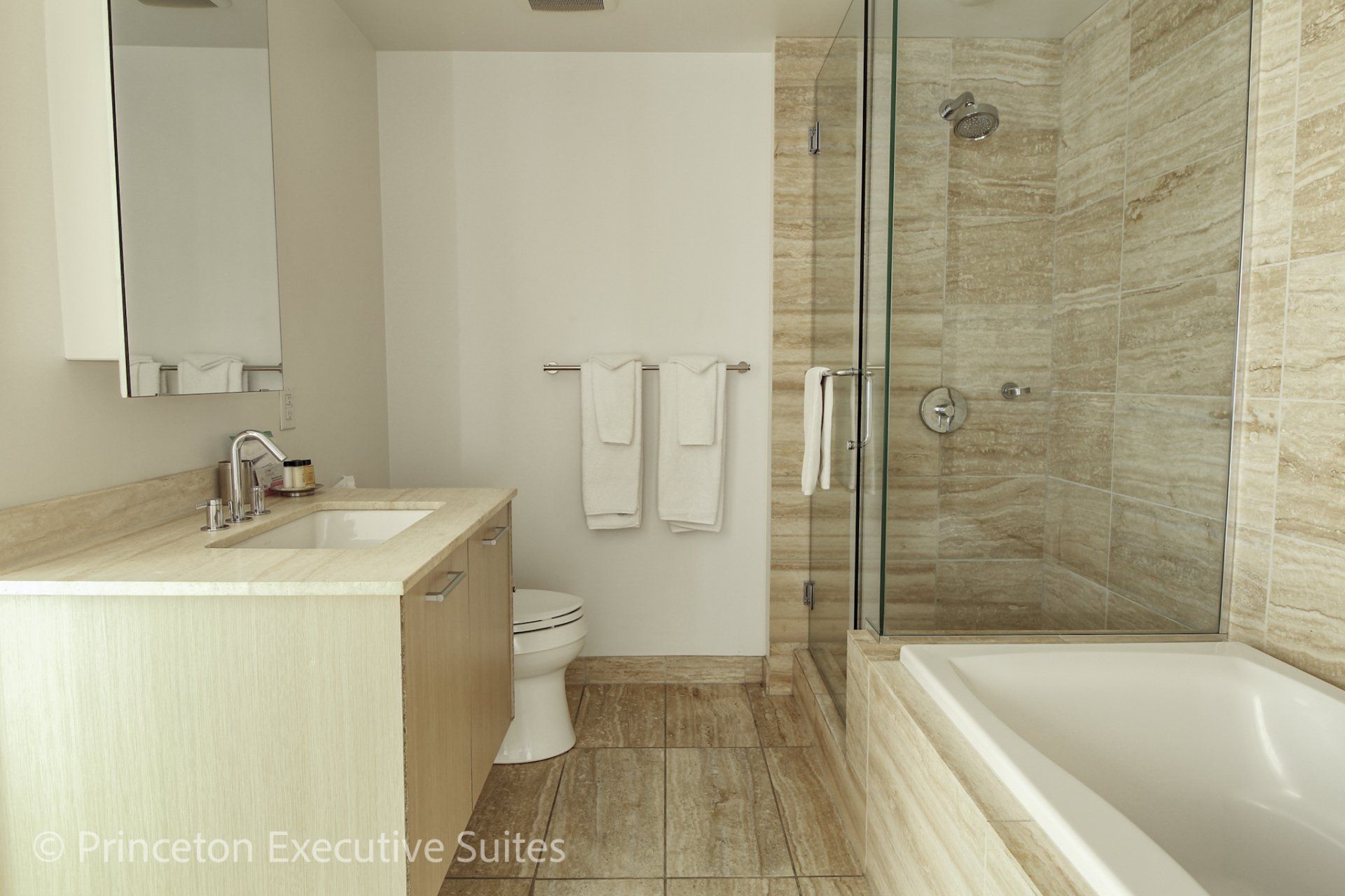 Bathroom with tan tiling, white tub, glass shower, and wooden vanity.