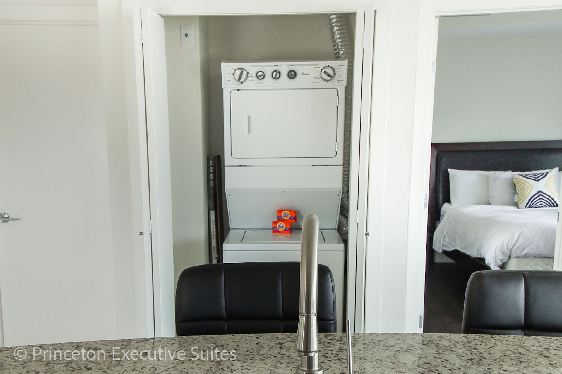 Stacked washer and dryer unit in a small laundry closet, open to a bedroom with a bed.
