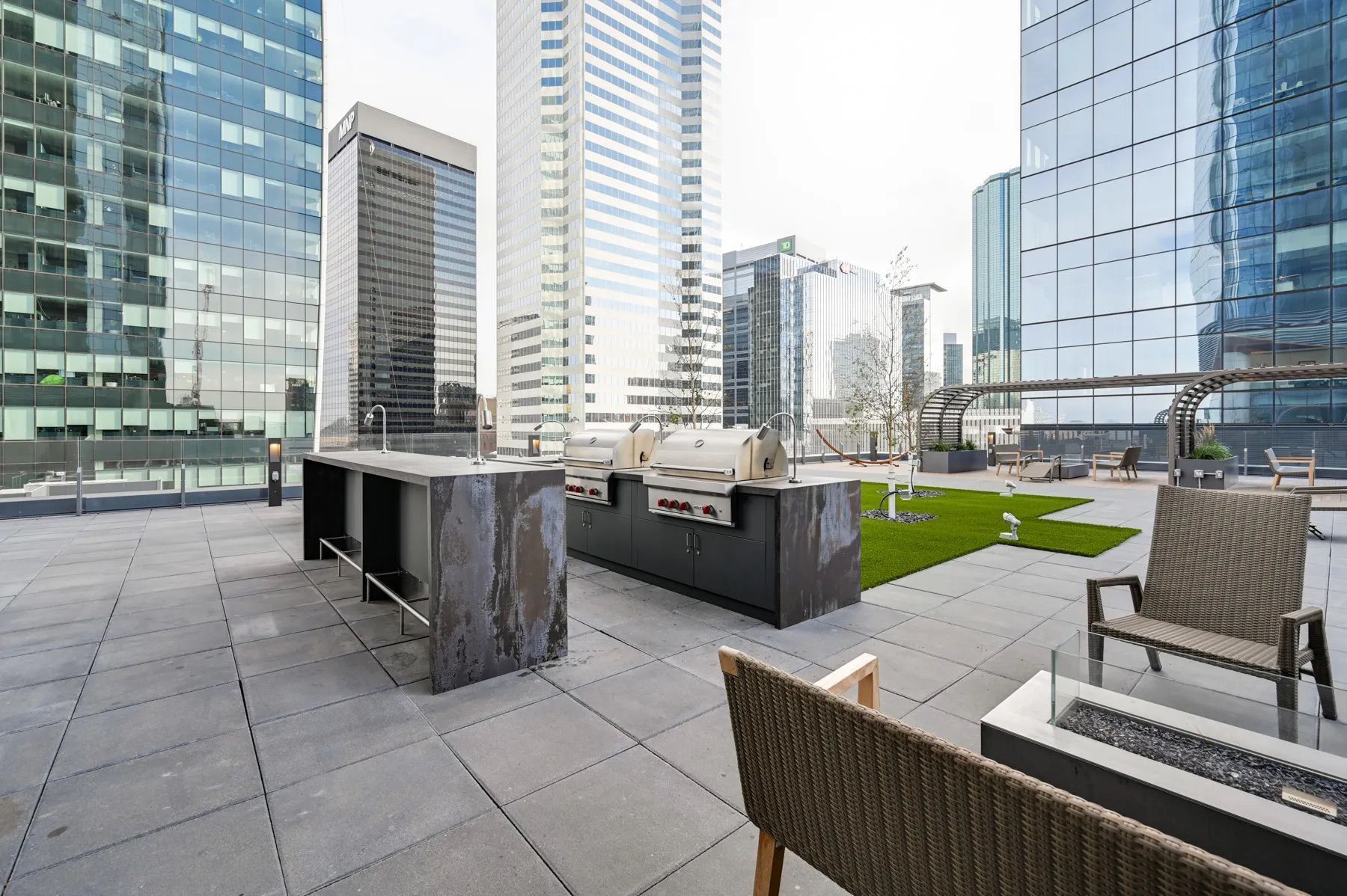 Rooftop patio with seating, grills, and cityscape backdrop. Modern high-rise buildings with reflective glass.