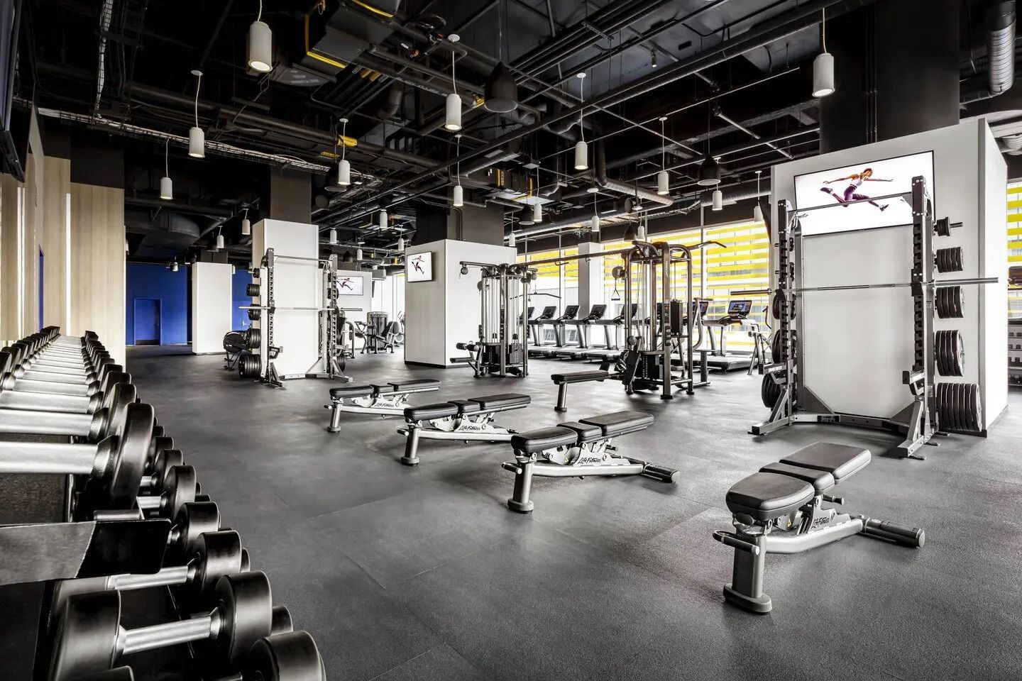Gym interior with weight benches, free weights, and exercise machines on a black rubber floor.