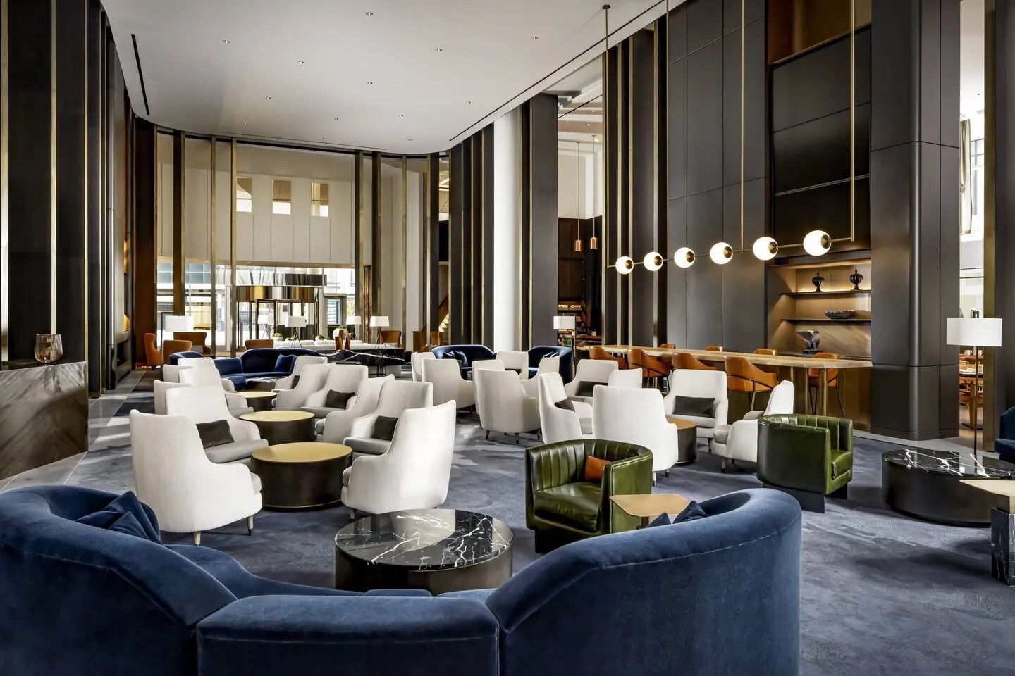 Spacious hotel lobby with modern furniture: blue couches, white chairs, dark walls, and long light fixtures.