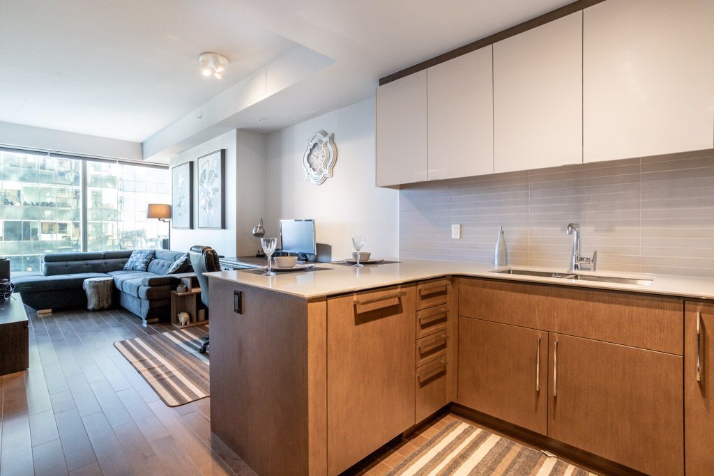 Modern kitchen with wood cabinets, island, and living room with dark sofa and large windows.