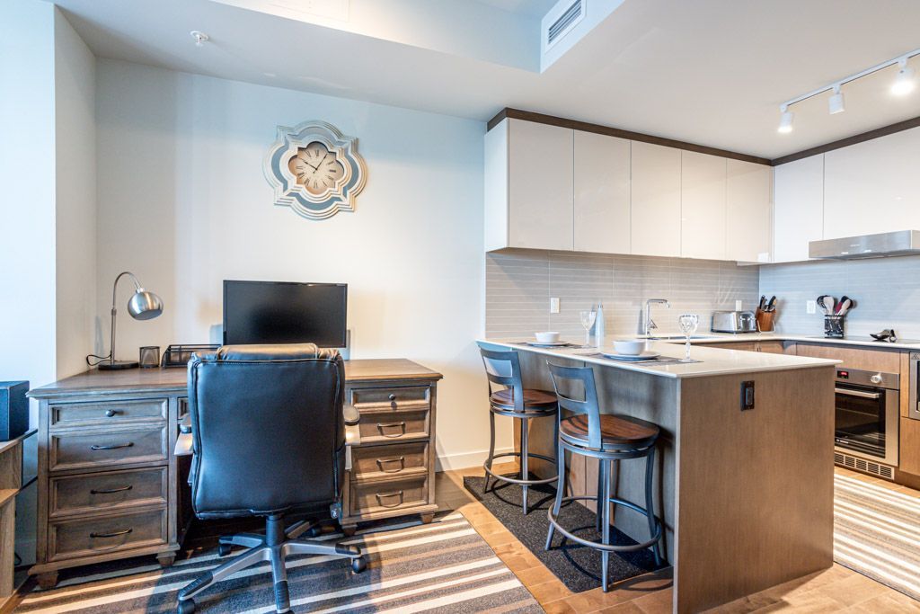 Modern apartment interior with desk, kitchen island with bar stools, and decorative wall clock.