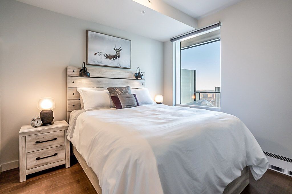 Bedroom with a white bed, wooden headboard, nightstand, and large window with a city view.