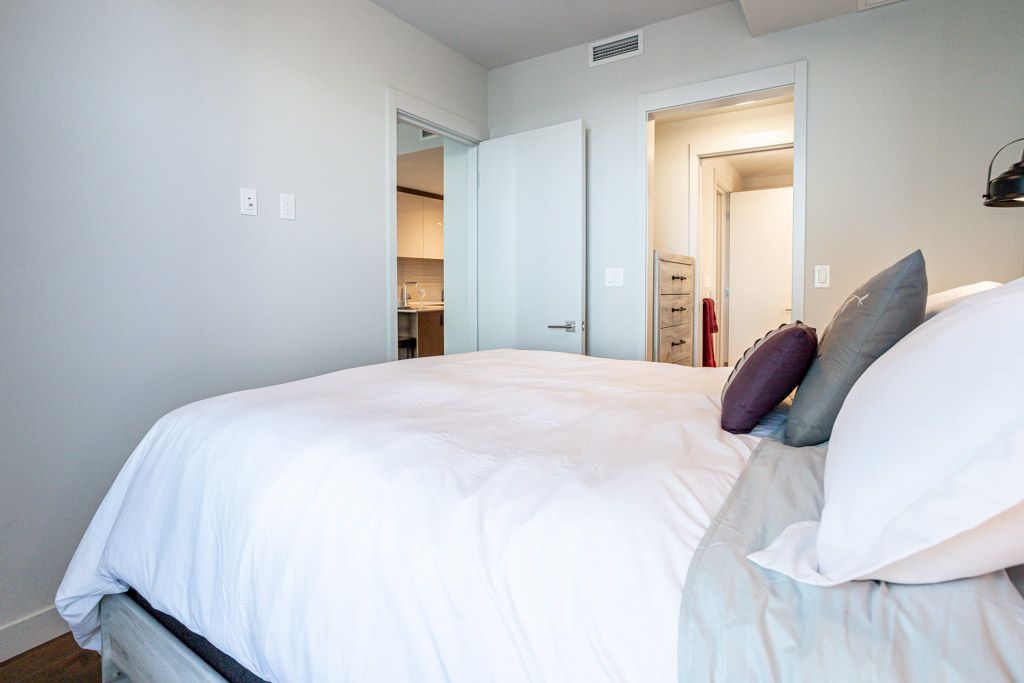 Bedroom with white bedding, gray wall, and open doors to kitchen and bathroom.