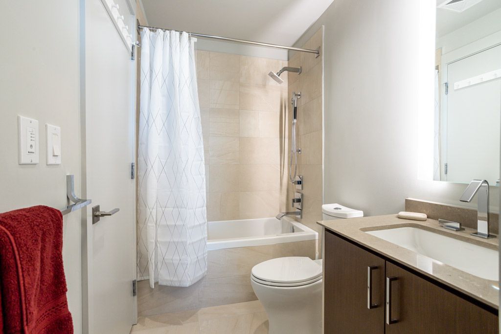 Small modern bathroom with a shower/tub, sink, and toilet. Red towel on the door.