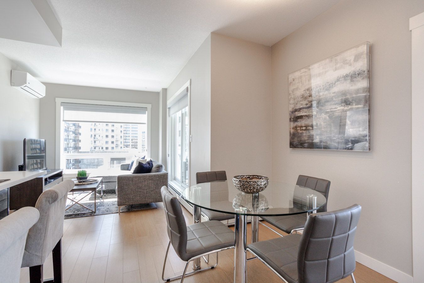 Living and dining room with glass table and grey chairs, large window, neutral colors, and artwork.
