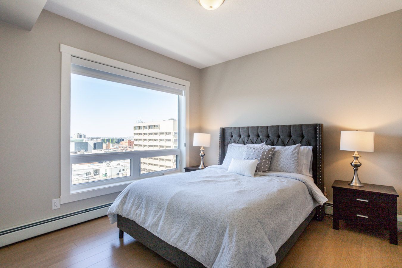 Bedroom with gray bed, large window with city view, and bedside lamps.