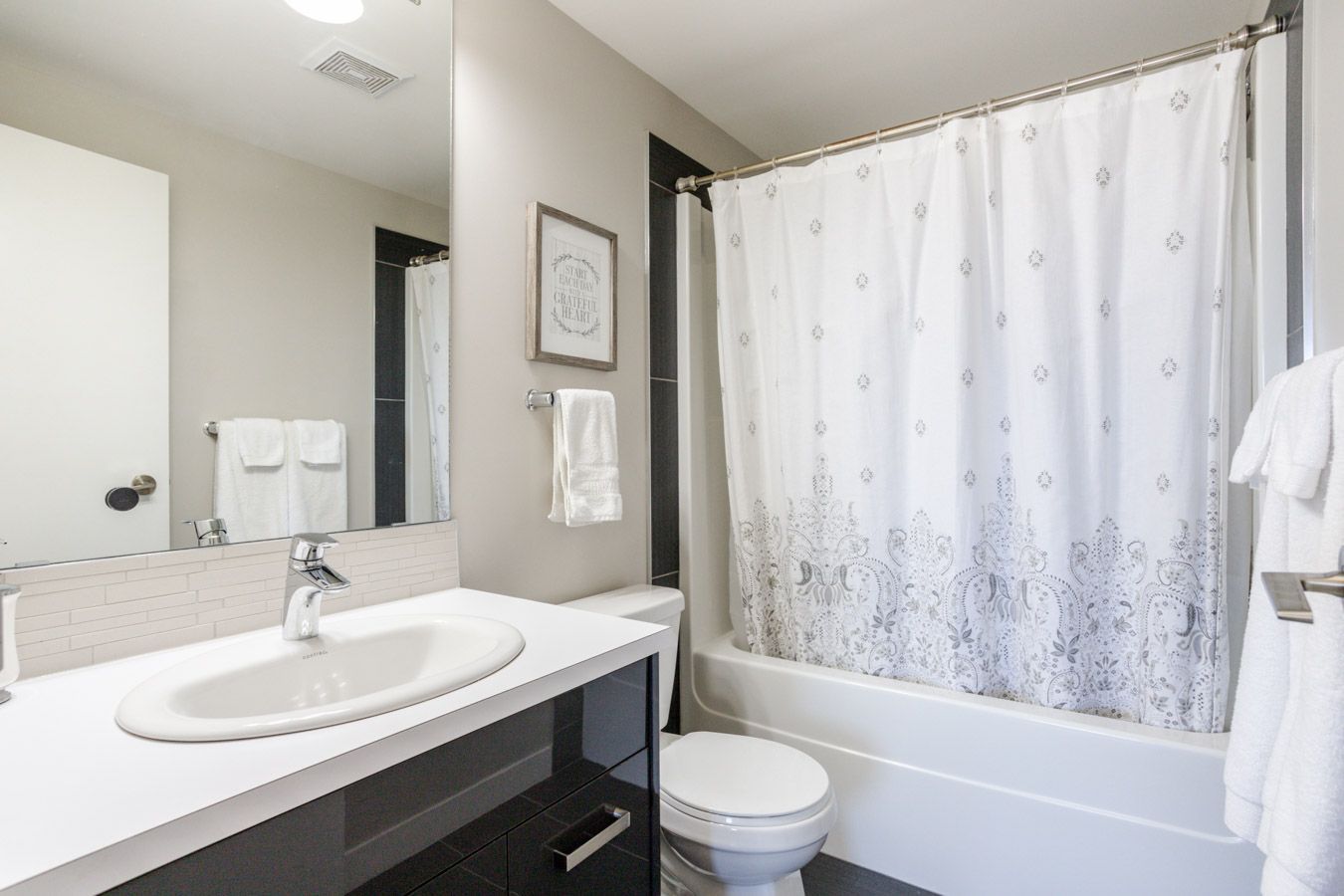 Modern bathroom with white sink, tub, and toilet; gray walls; black accents; patterned shower curtain; and white towels.