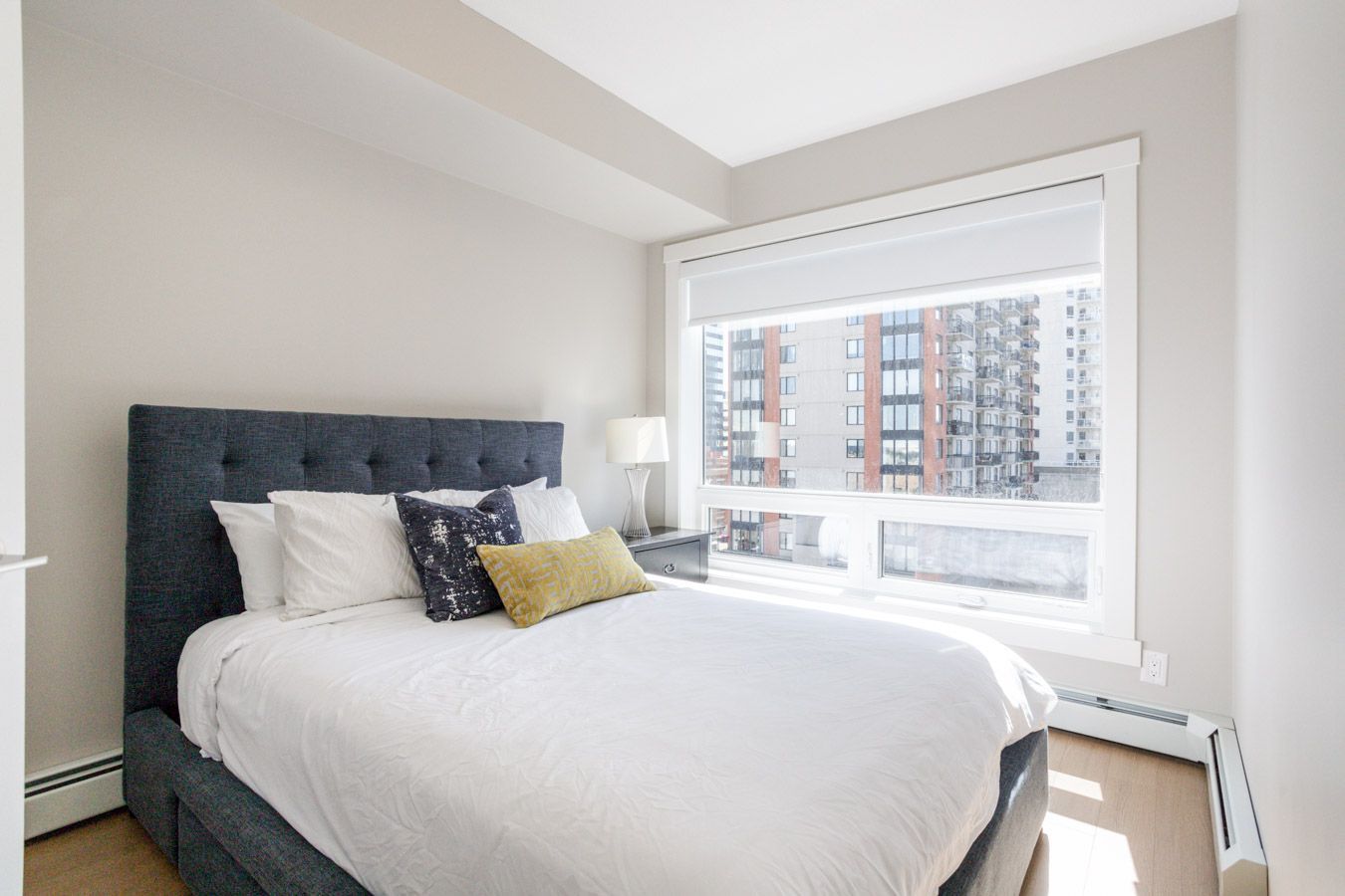 Bedroom with a bed against the wall, large window, and bedside table. Neutral color scheme with pops of blue and yellow.