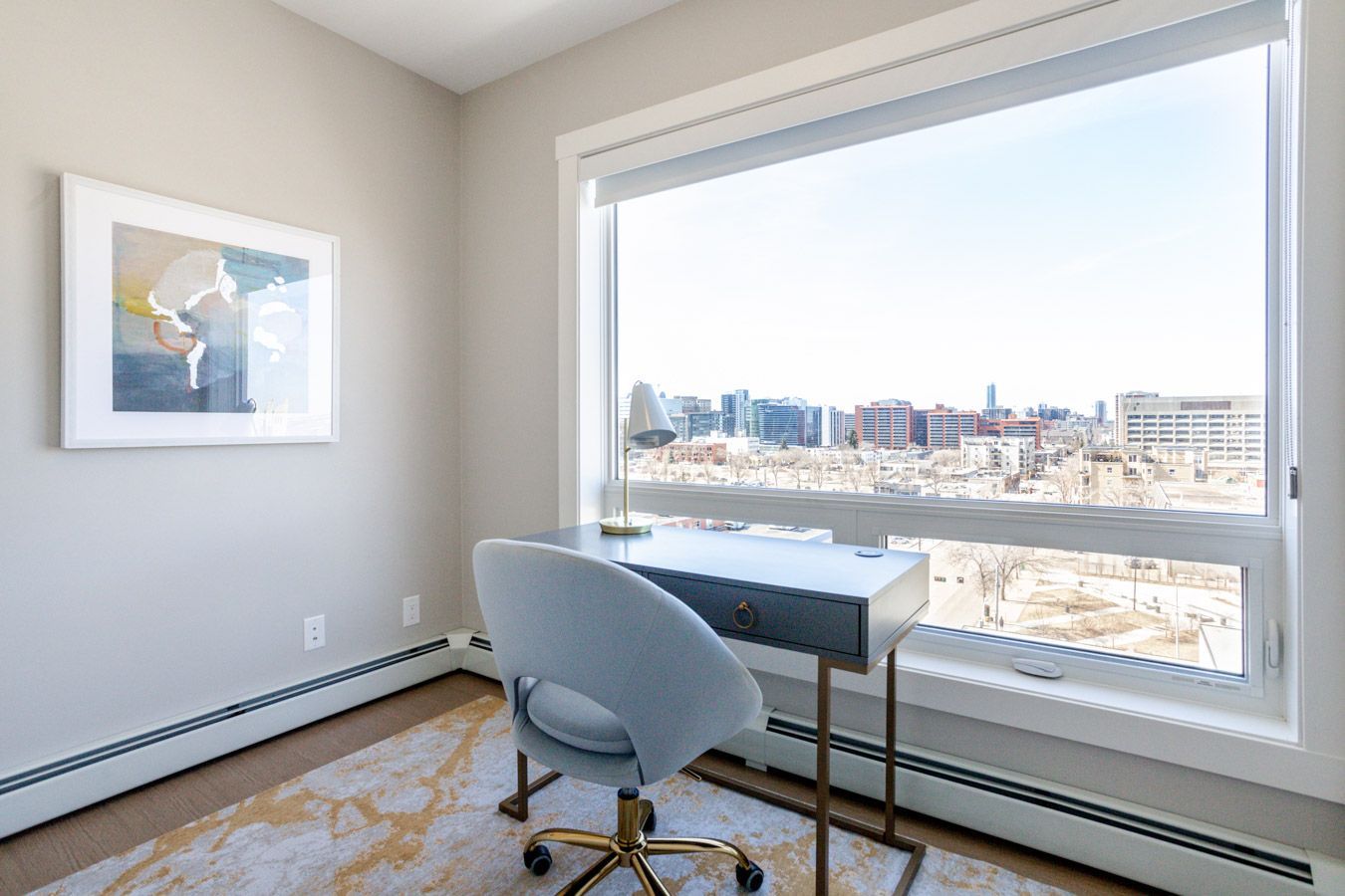 Desk with chair near a large window overlooking a city, neutral tones, art on wall.