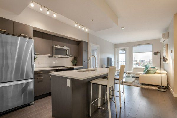 Modern condo kitchen and living room. Stainless steel appliances, island, white sofa, and dark wood floors.