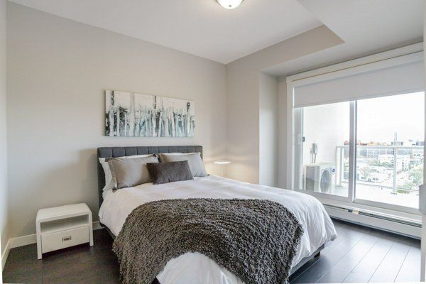 Bedroom with a bed, nightstand, and large window overlooking a city view. Gray walls, dark wood floor.