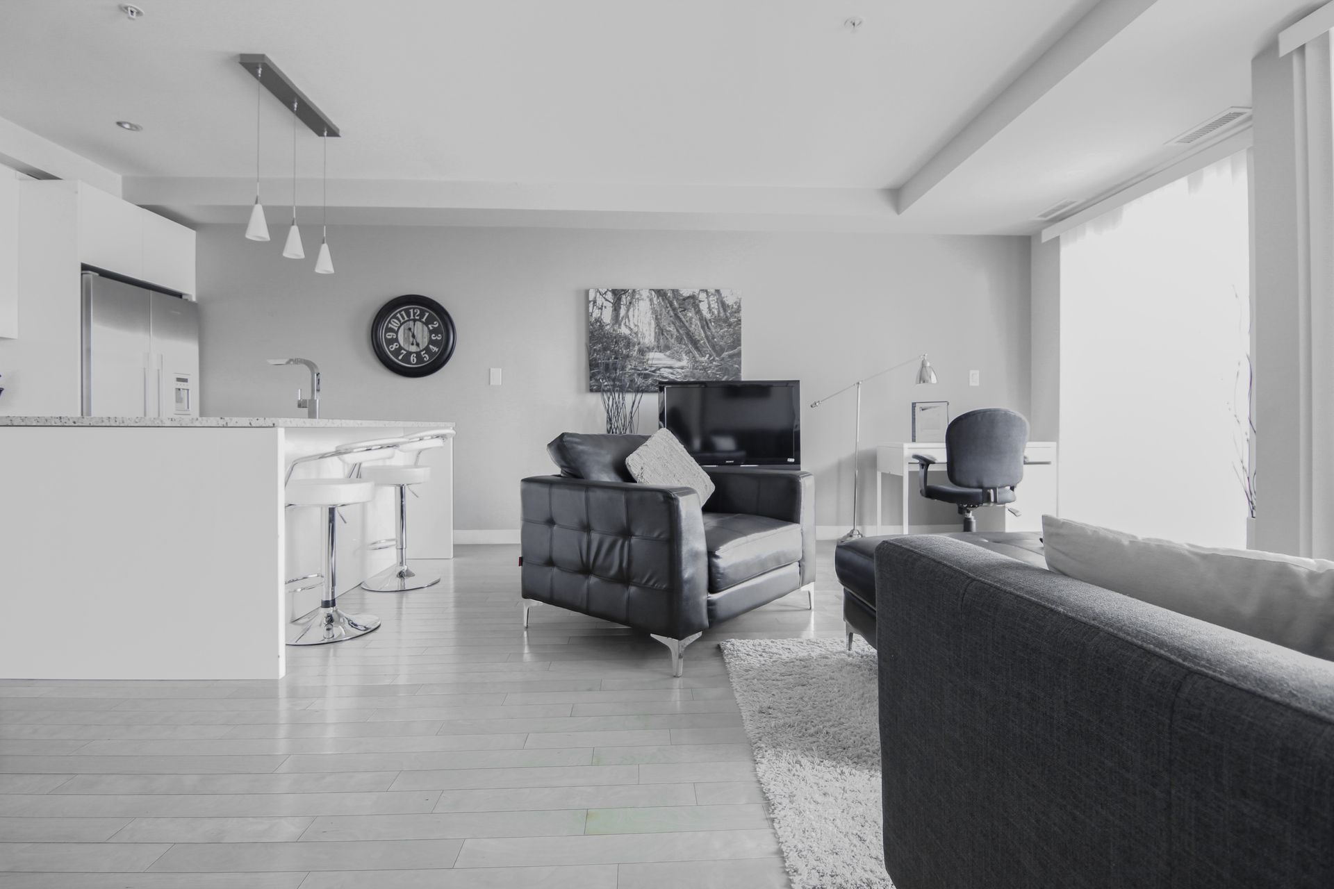 Modern, open-concept living space in grayscale. Kitchen island with stools, sofa, armchair, desk, large windows.