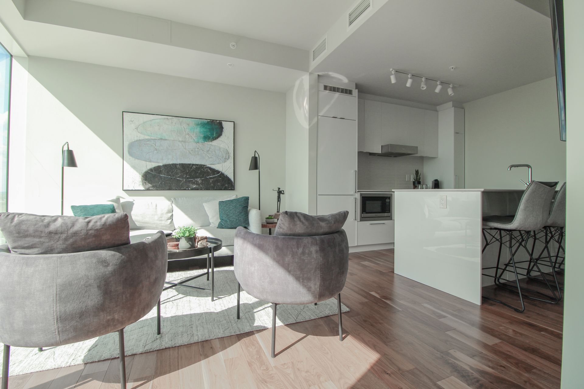 Modern living room with chairs, sofa, kitchen, and art. Light wood floors, white walls, and lots of natural light.