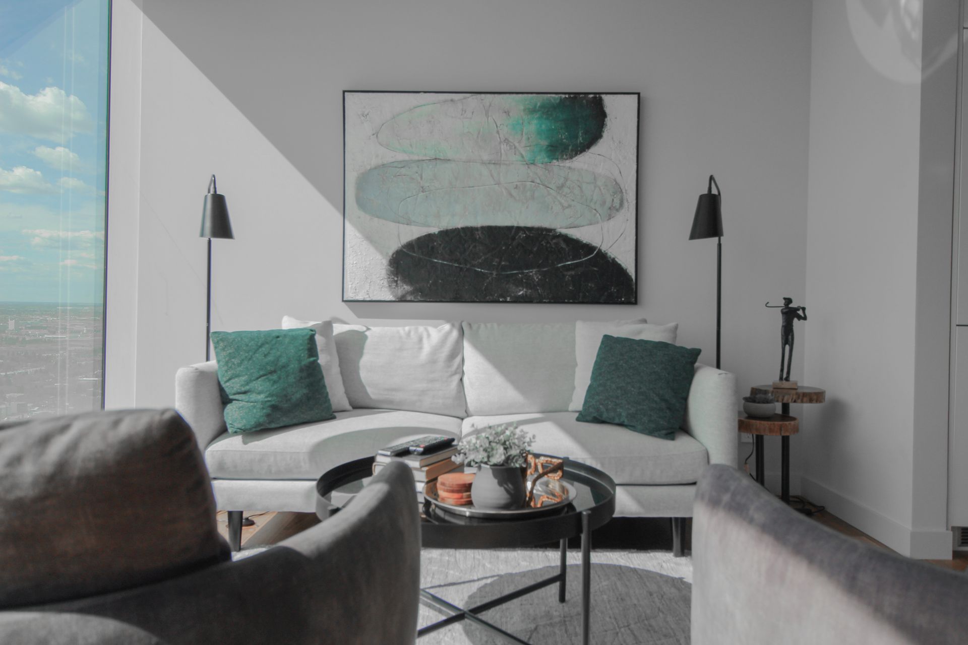 Living room with white sofa, teal pillows, art, and two black lamps, with cityscape view.