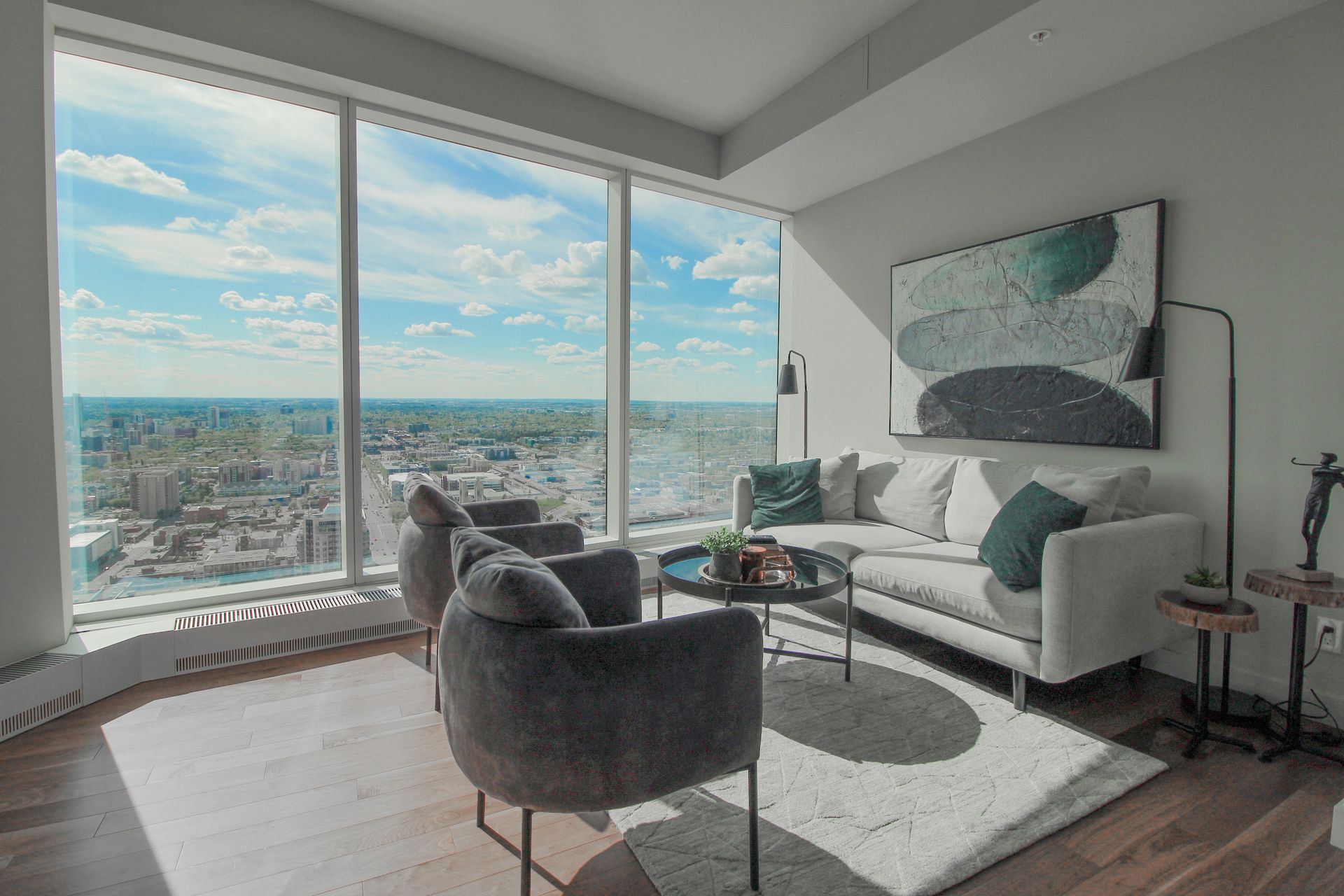 Living room with large windows overlooking a city. Two gray armchairs, a white couch, and a rug are present.