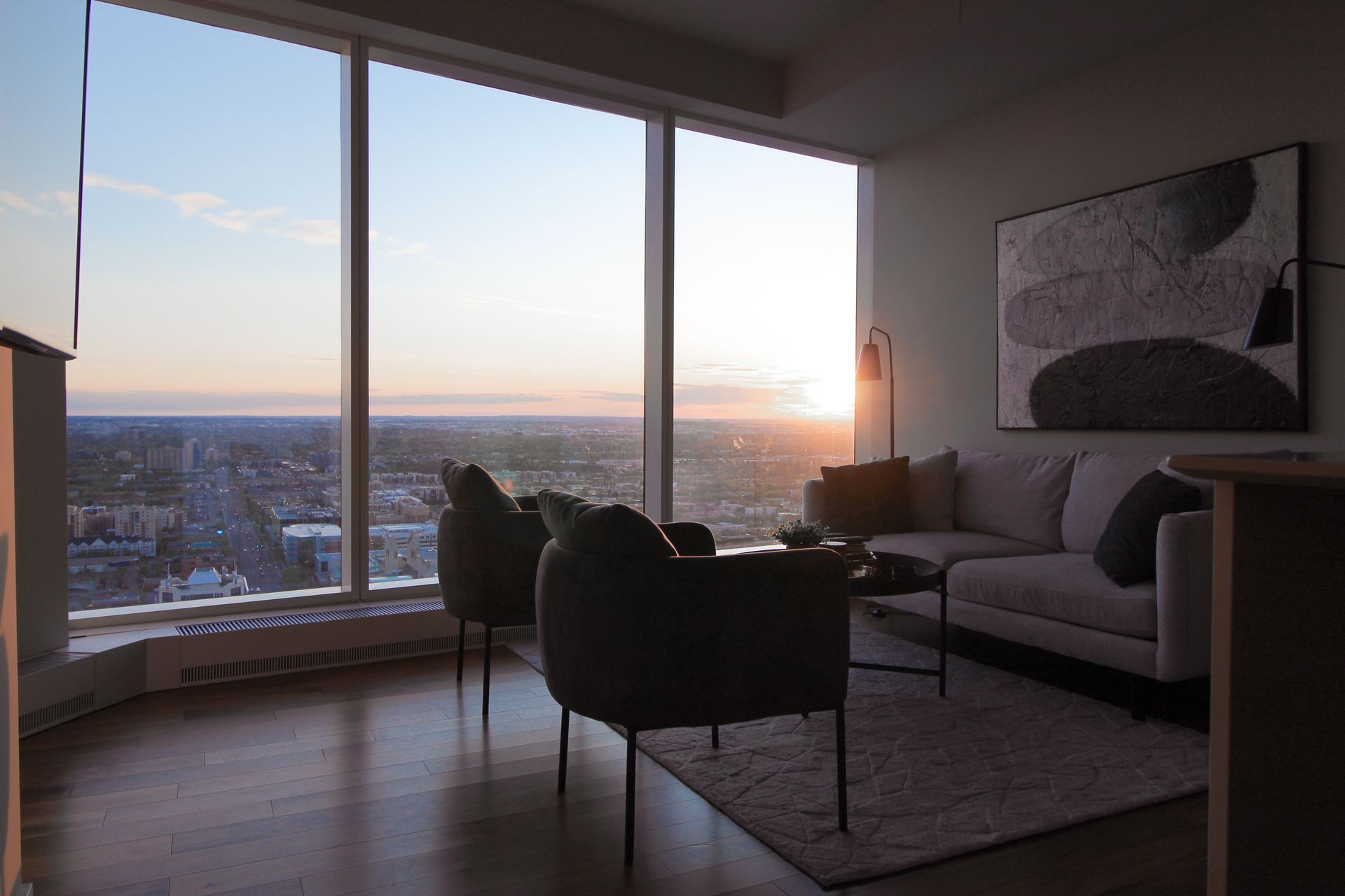 Living room with large windows, cityscape view at sunset. Two chairs face sofa, art on wall.