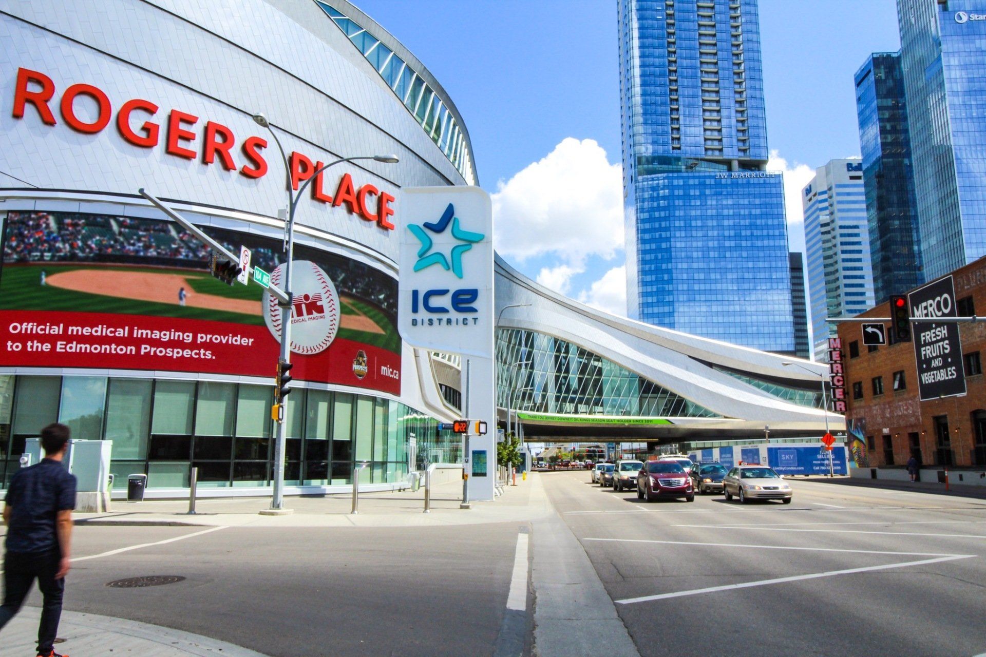 Rogers Place arena exterior on a sunny day, street in the foreground with cars, person walking.