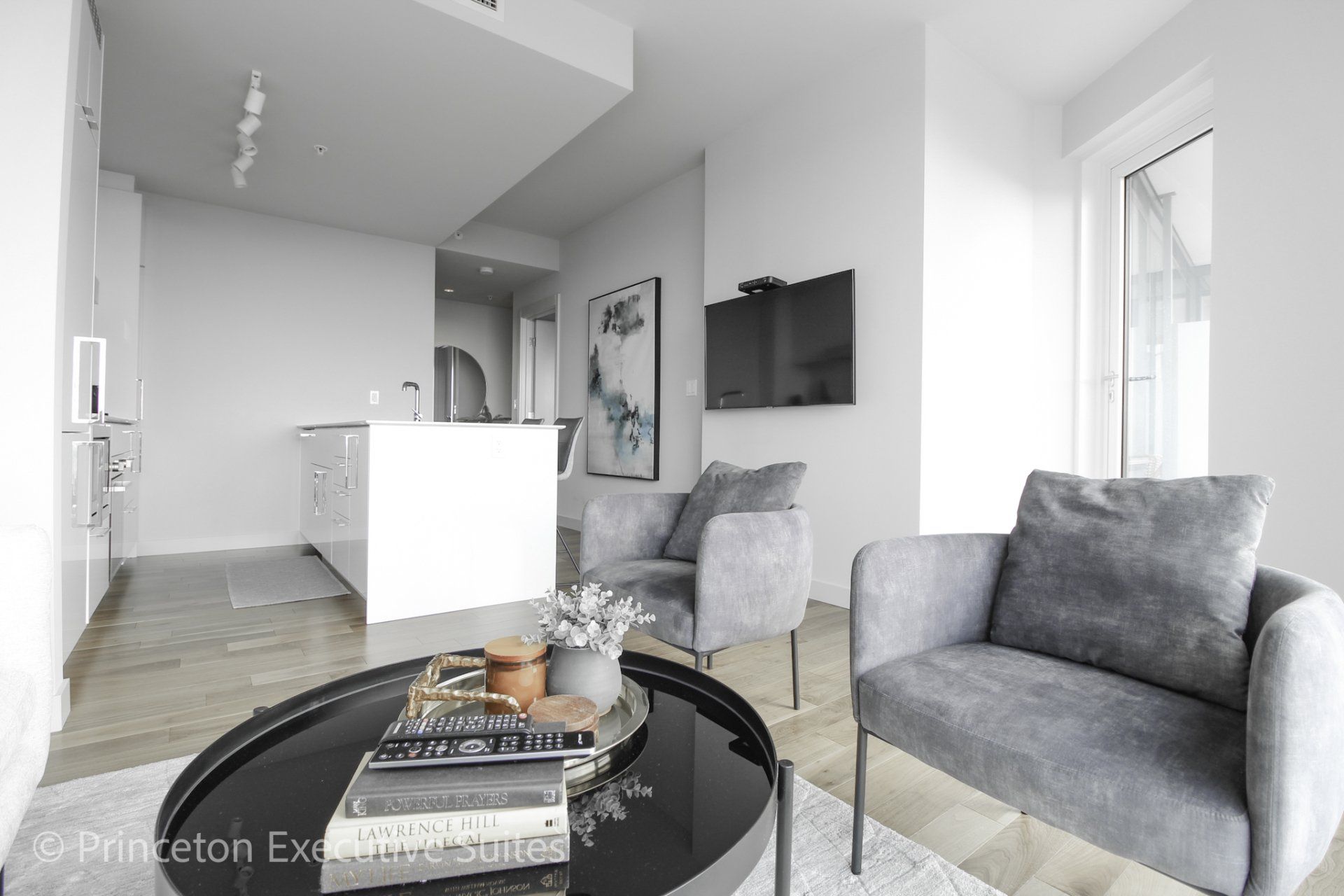 Open concept living area in this edmonton furnished suite with white kitchen Grey accent chairs round glass coffee table.