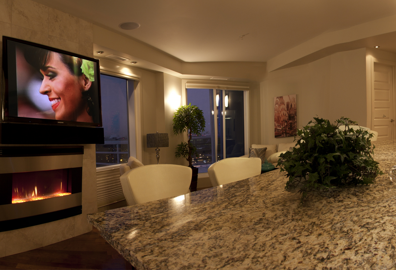 Cozy living room with TV over fireplace. Granite countertop, chairs. City view through windows.