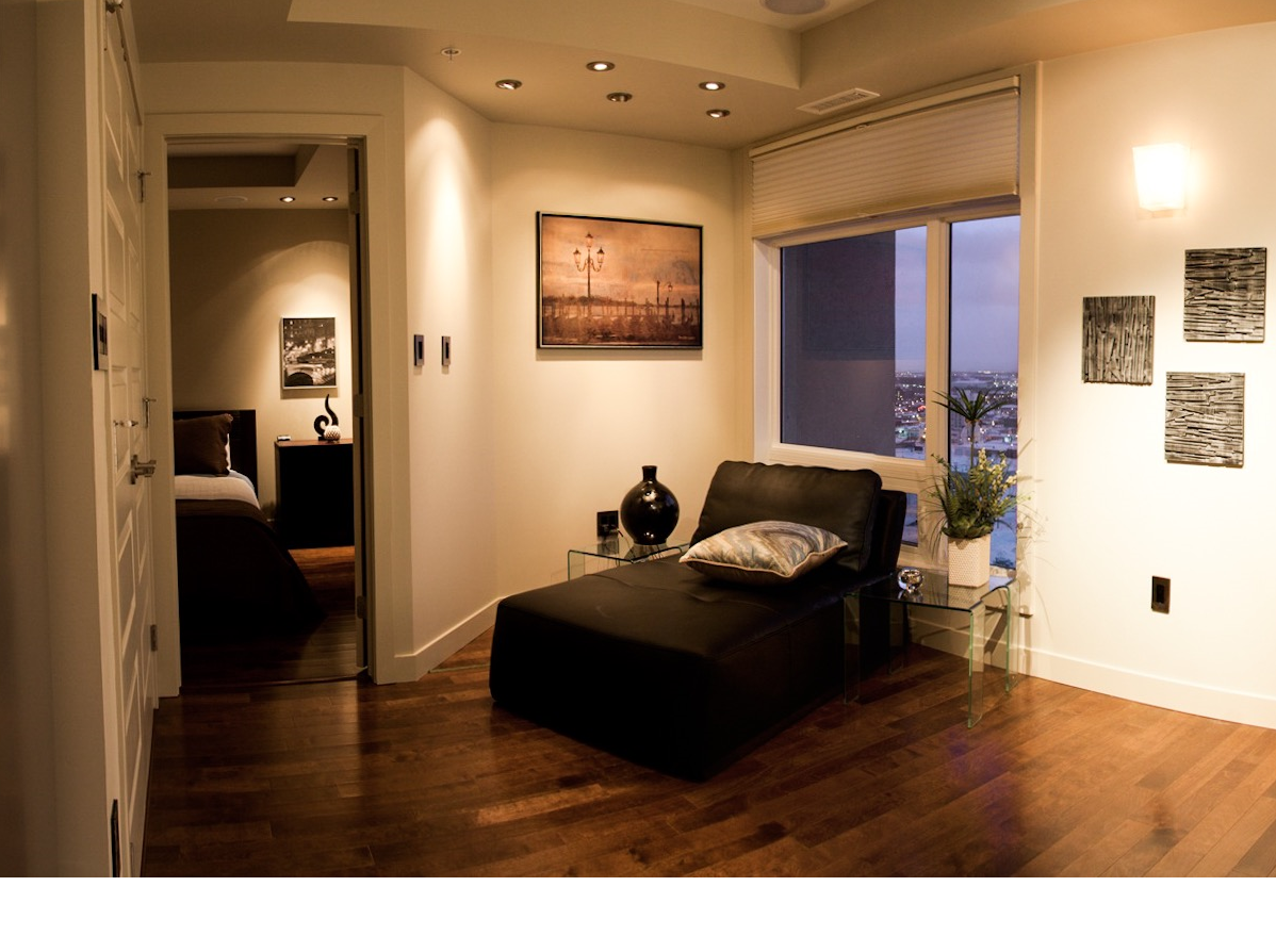 A modern bedroom with a chaise lounge, artwork, and a view. Warm lighting and hardwood floors.