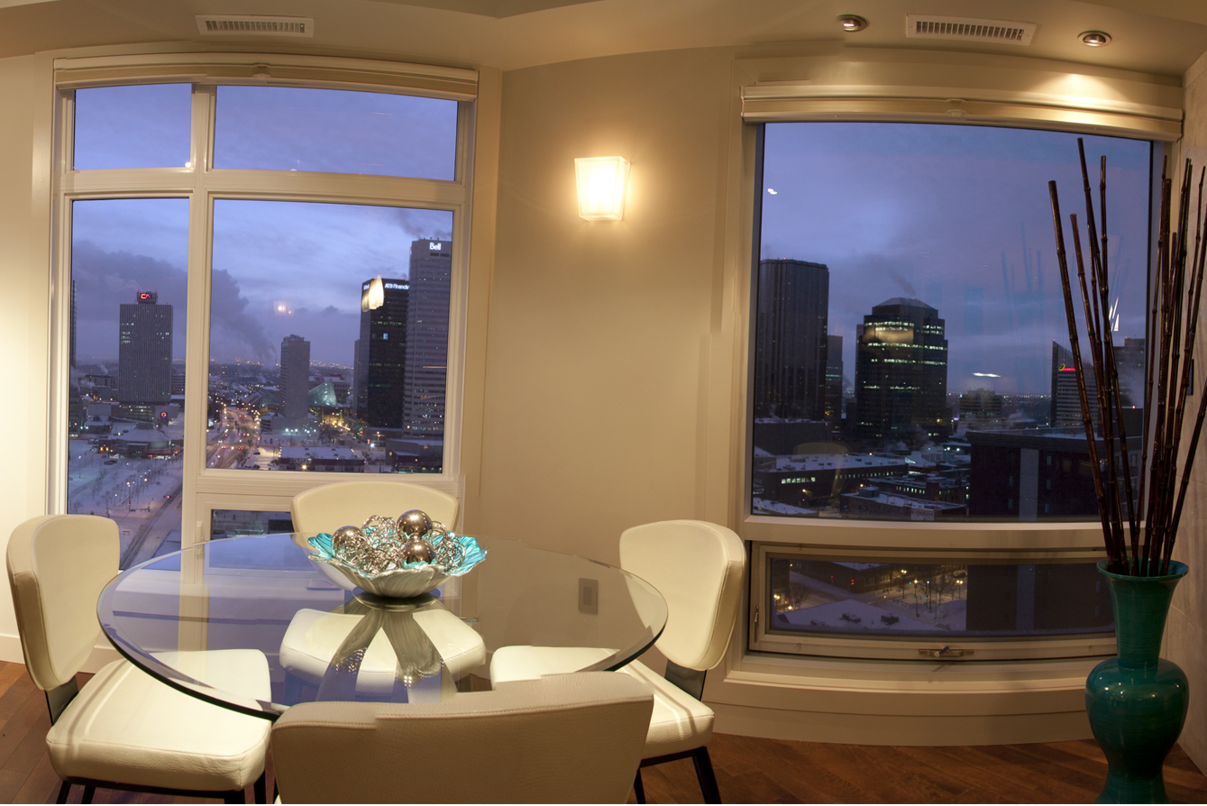 Modern dining room with city view at dusk. Round glass table, white chairs, and decorative vase.