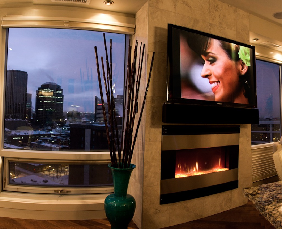 Cozy room with cityscape view; TV above fireplace. Woman smiling on TV. Green vase with brown sticks.