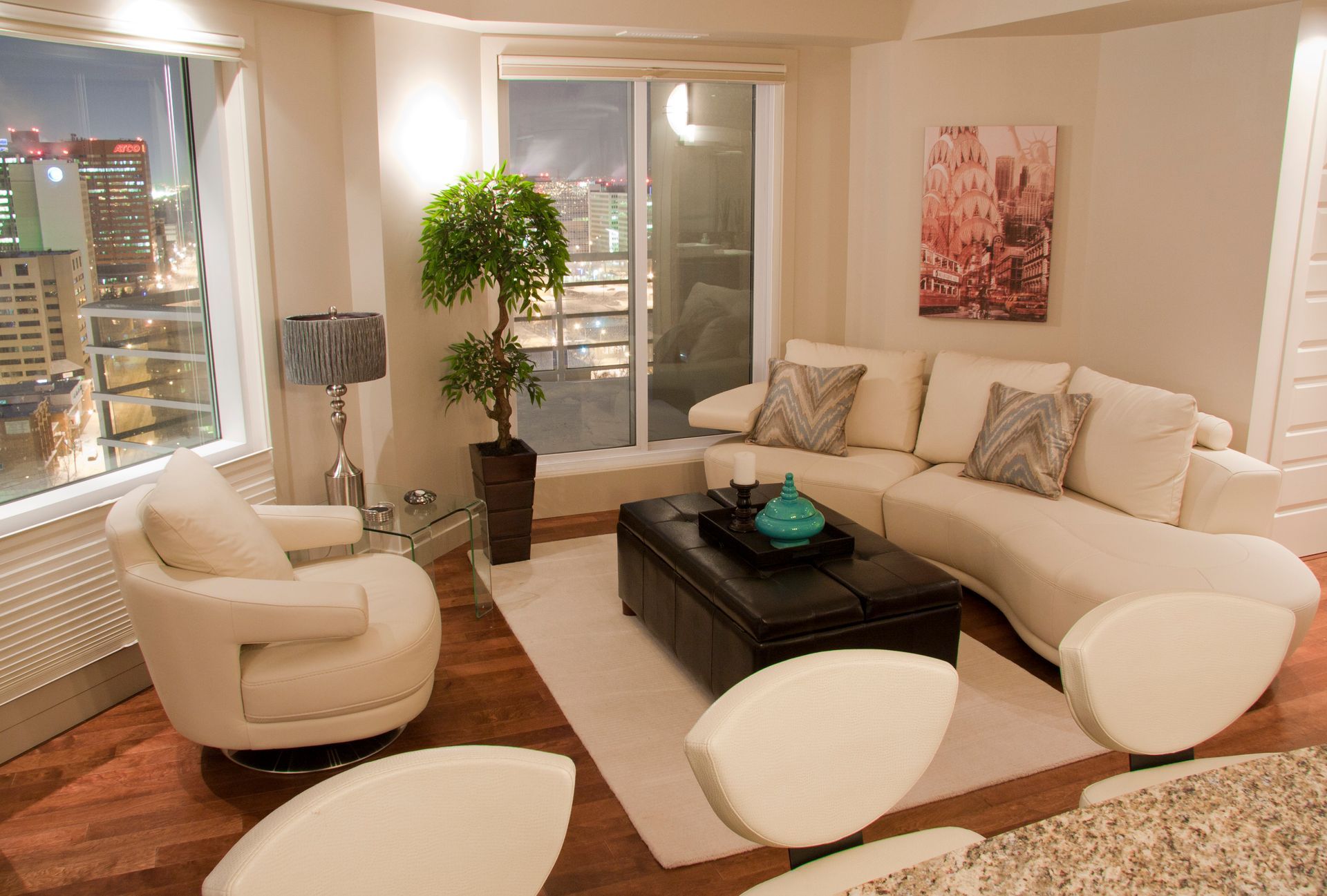 Living room with white sofa, chairs, and rug; city view through windows.