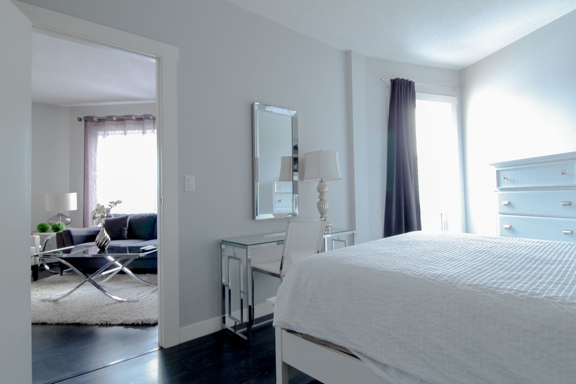 Bedroom with white bed, mirrored vanity, and view into living room. Light walls and dark floor.