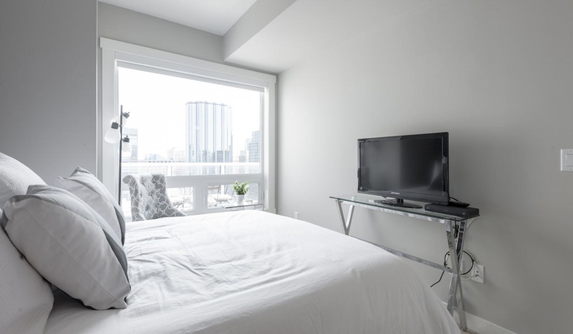 Bedroom with white bedding, large window, TV on a stand, and cityscape view.