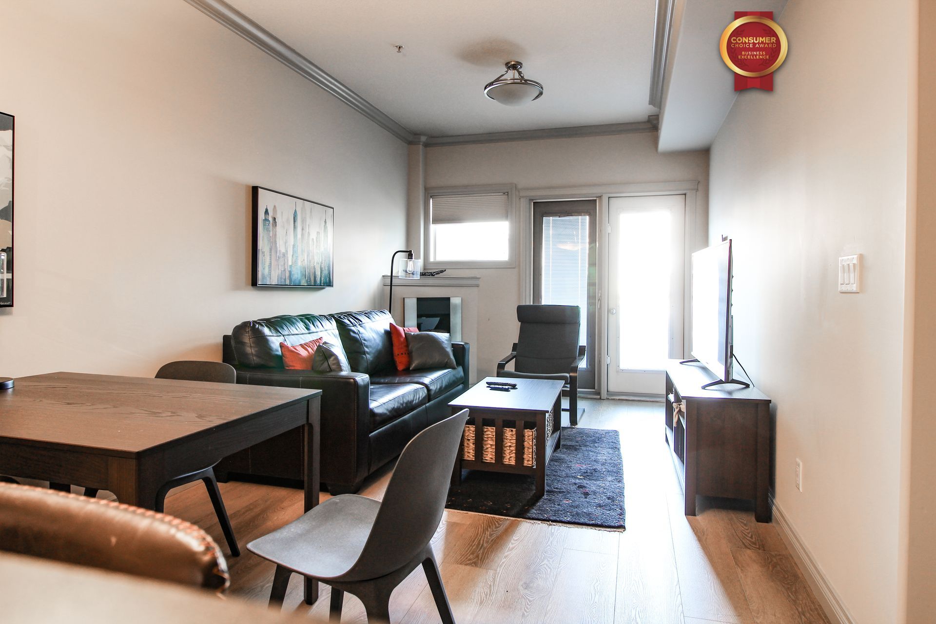 Living room with black leather sofa, TV, and wooden table and chairs. Bright light comes from a door.