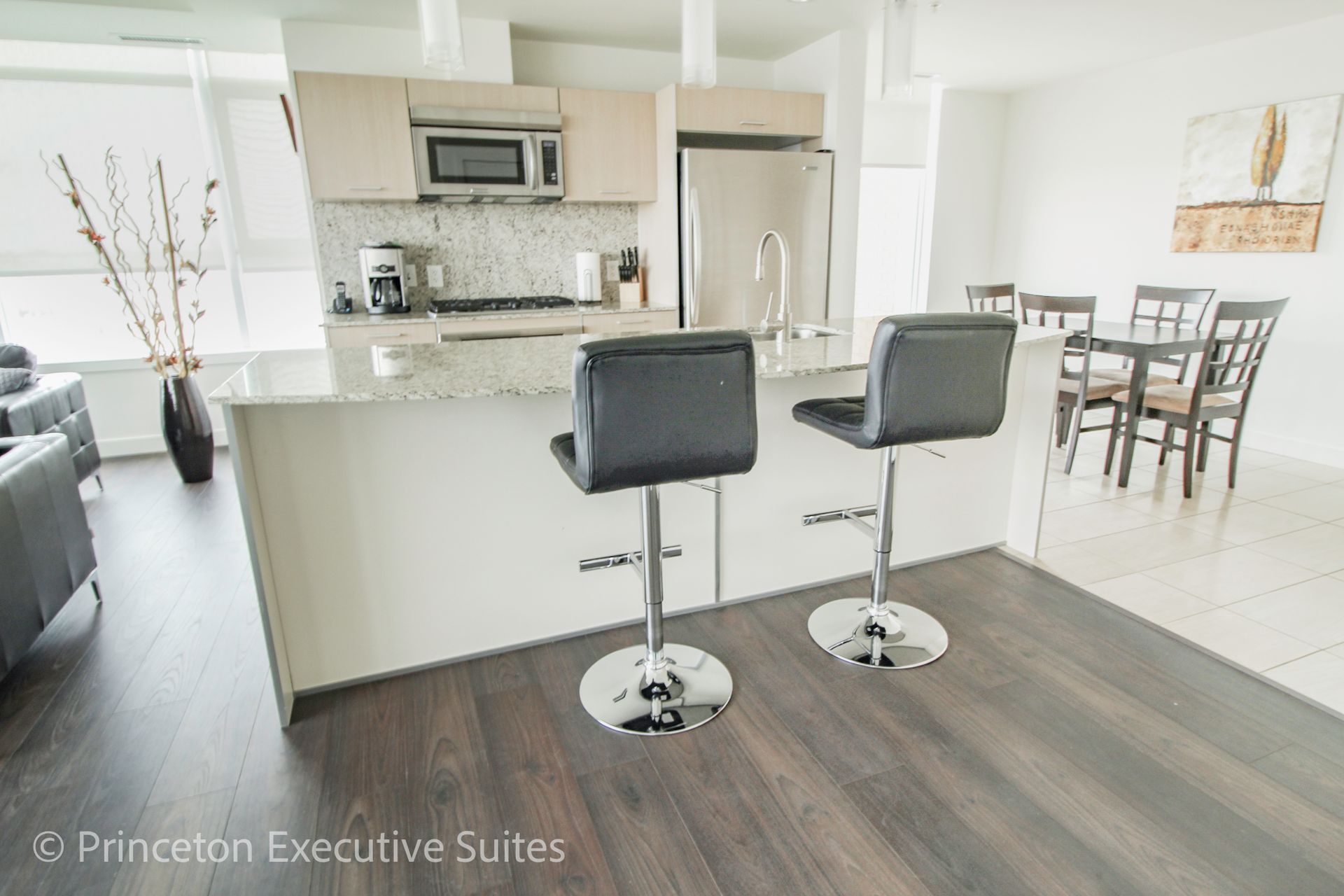 Modern kitchen with island, stools, and dining table. Dark wood floor, white walls, stainless steel appliances.