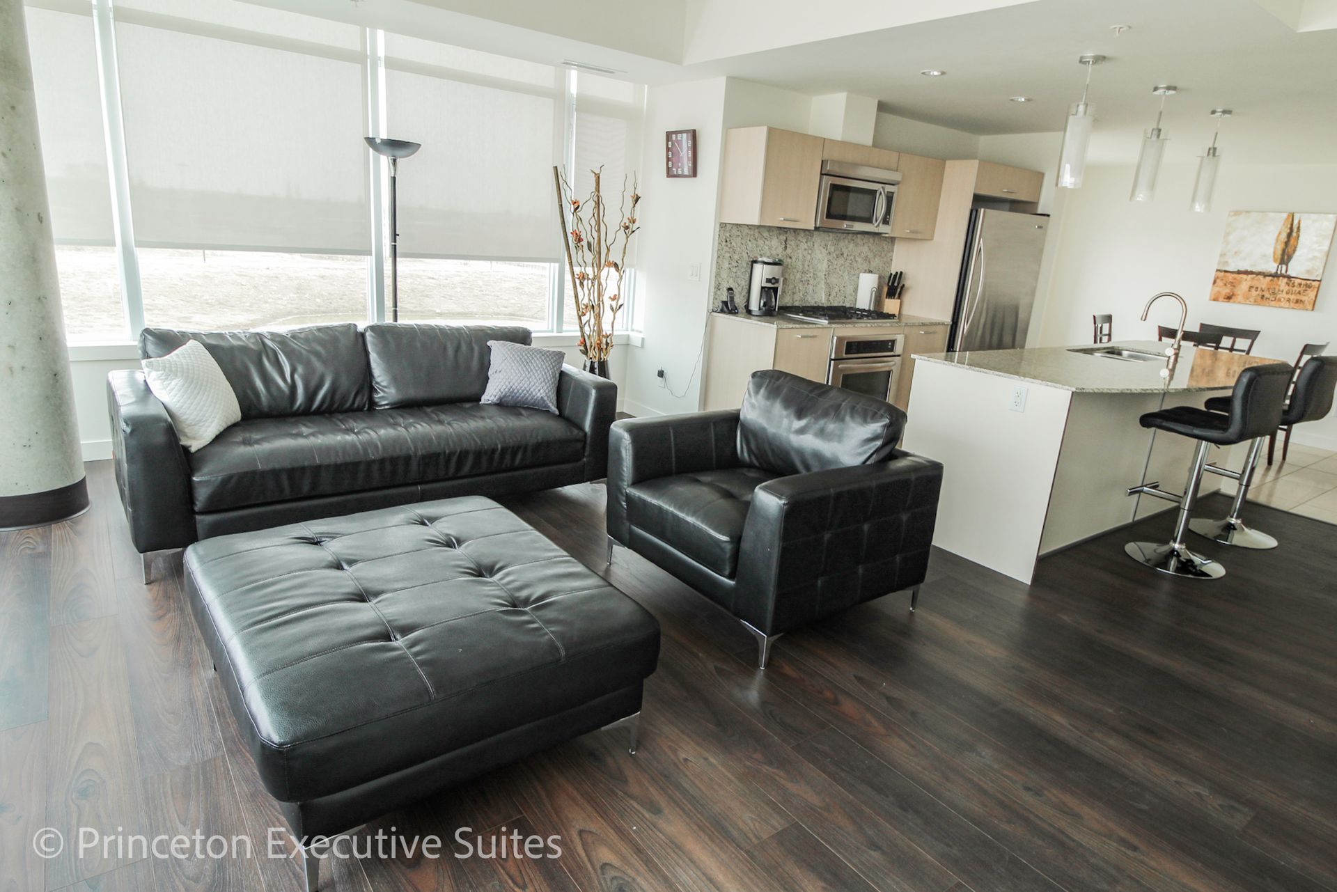Modern living room with black leather furniture, kitchen in background. Dark wood floors, large windows.