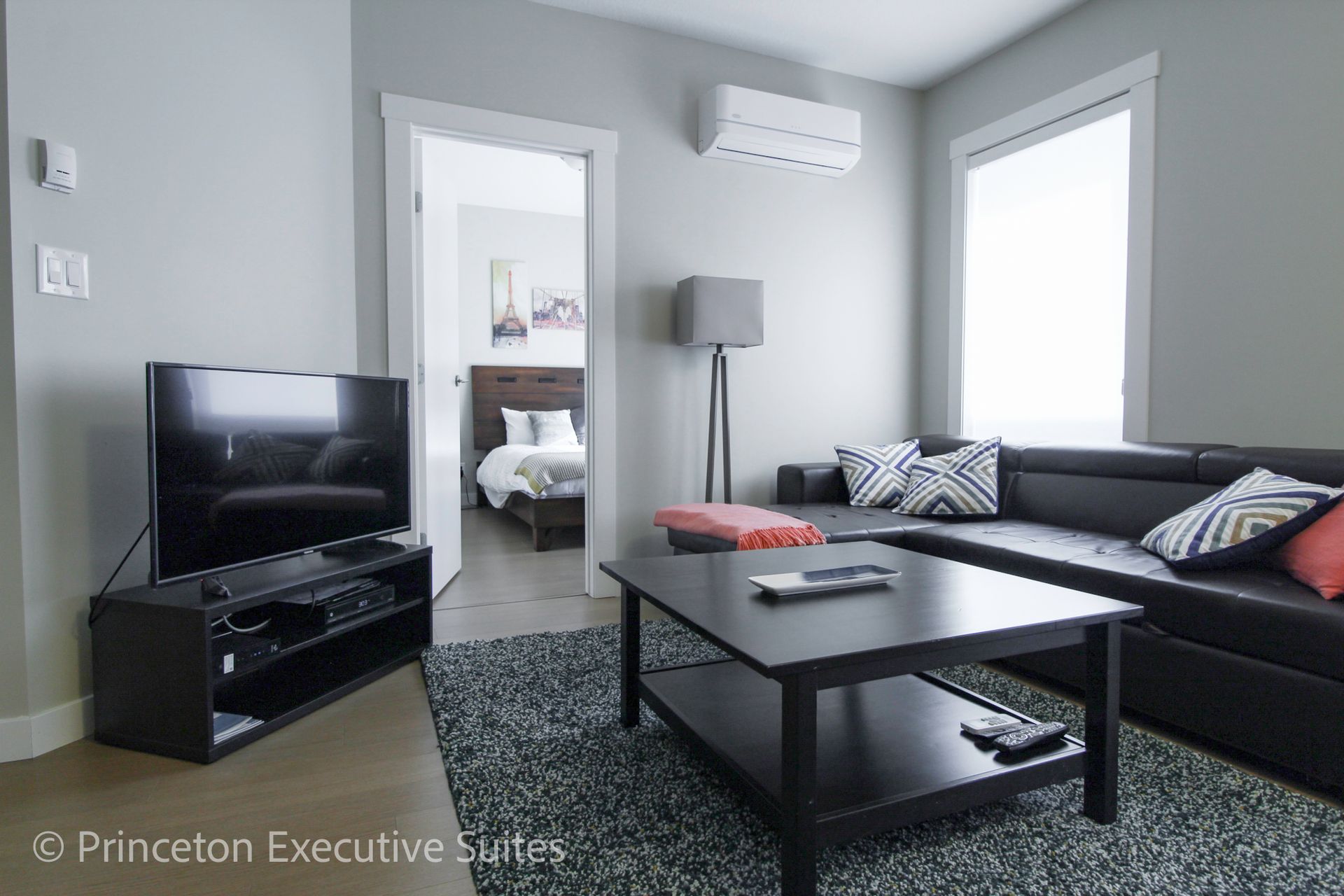 A modern living room with a black sectional, TV, and coffee table. A bedroom is visible in the doorway.