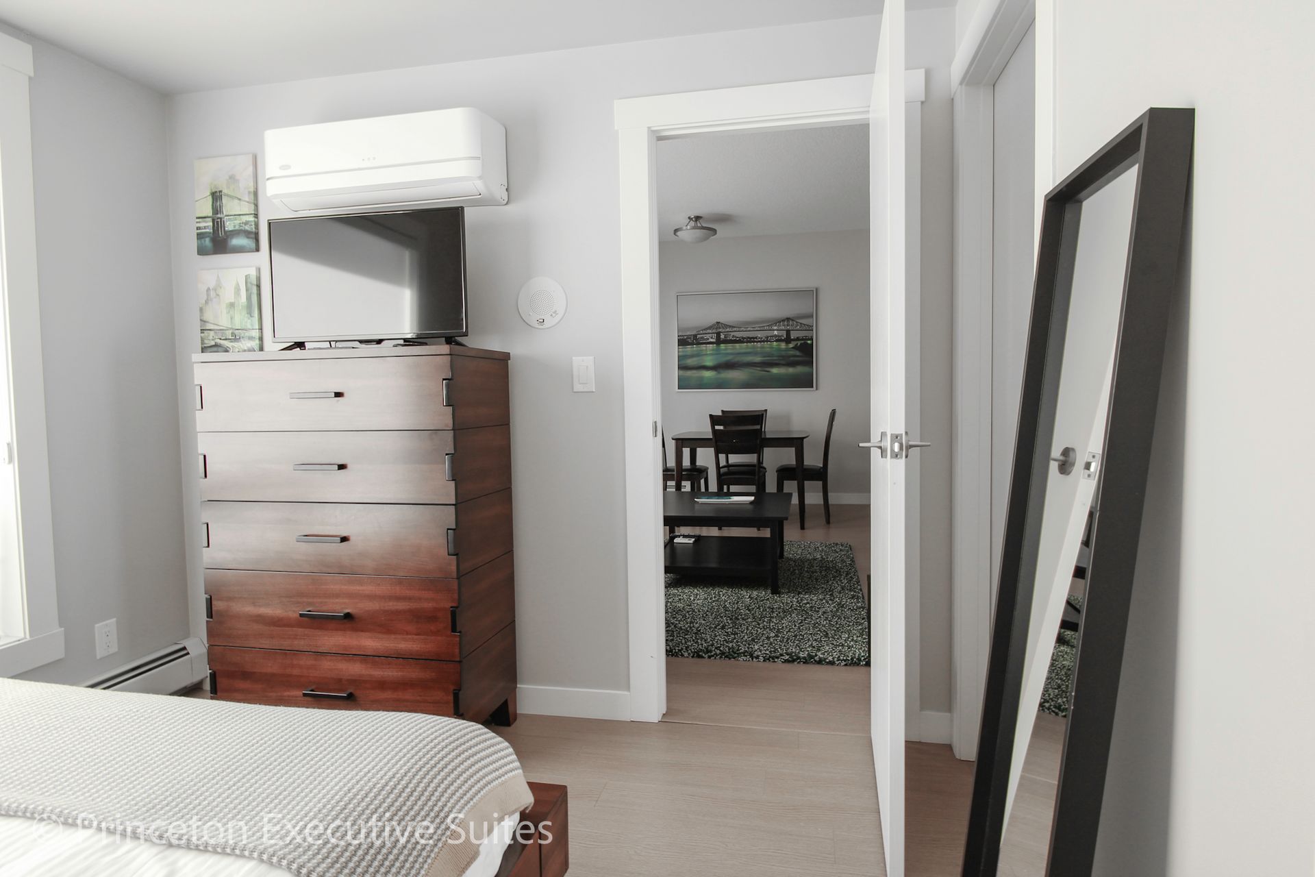 Bedroom with a wooden dresser, TV, and large mirror; a doorway leads to a dining area.