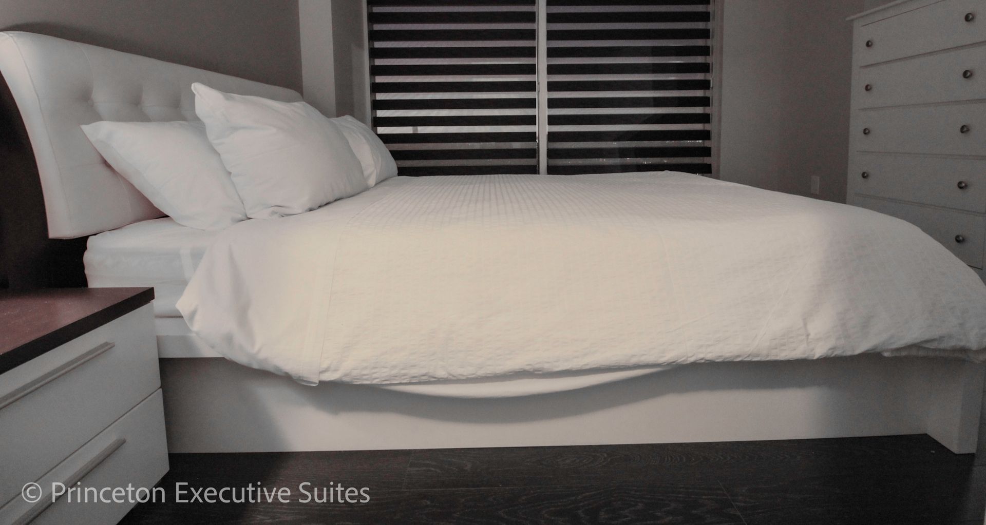 White-themed bedroom with a large bed, nightstand, and chest of drawers. Window with blinds in the background.