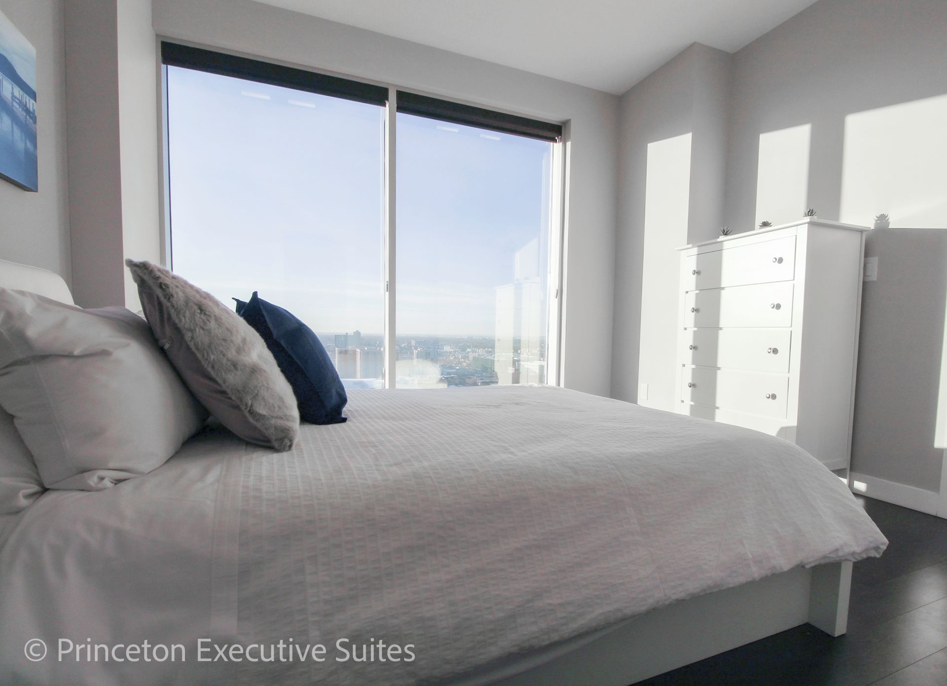 Bedroom with a bed, pillows, dresser, and large window. Sunlight streams in.