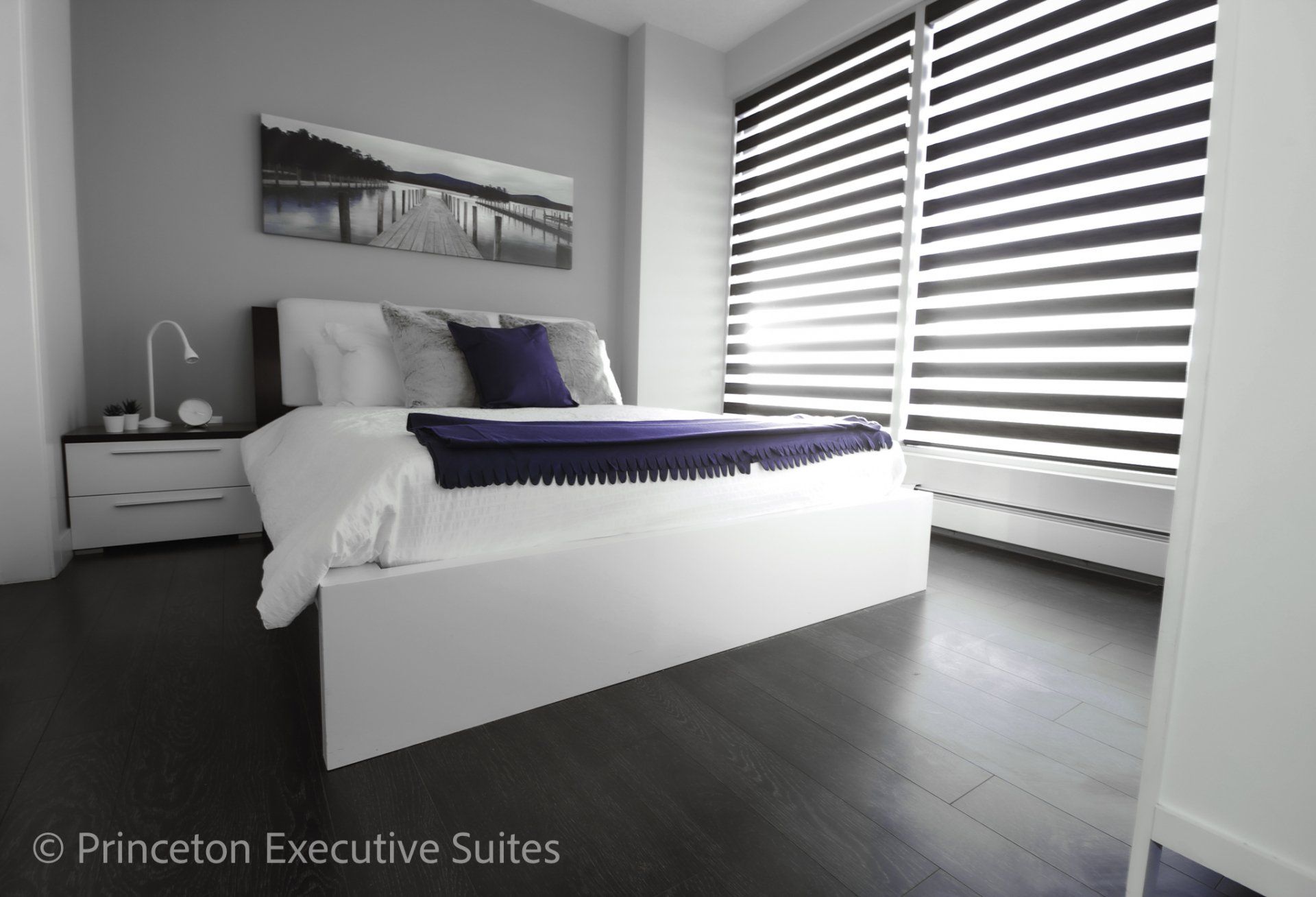 Modern bedroom with large bed, dark wood floors, gray walls, and window blinds.