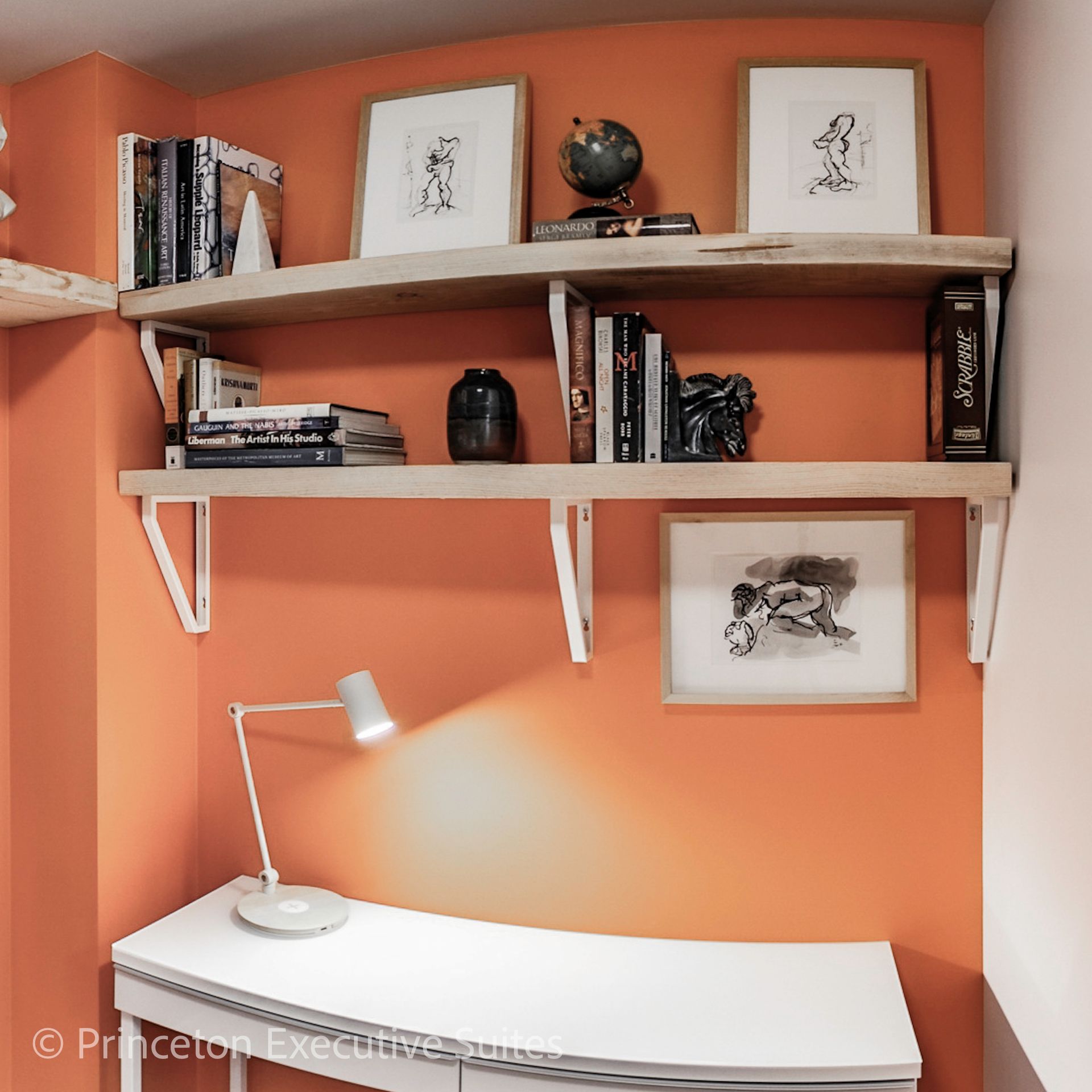 Orange-walled home office with white desk, bookshelves, artwork, and a lamp.
