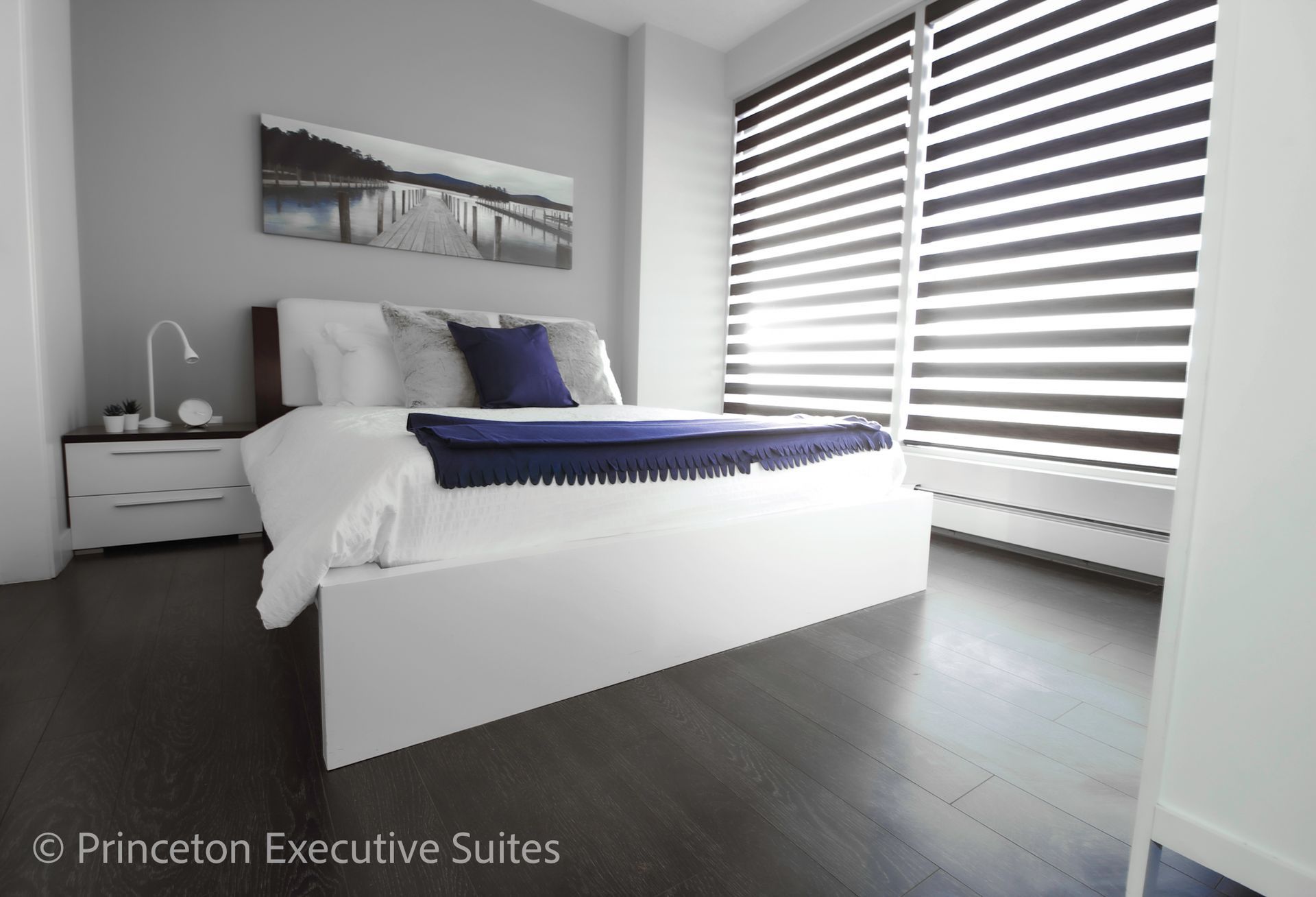 Modern bedroom with a white bed, gray walls, and large window with blinds.