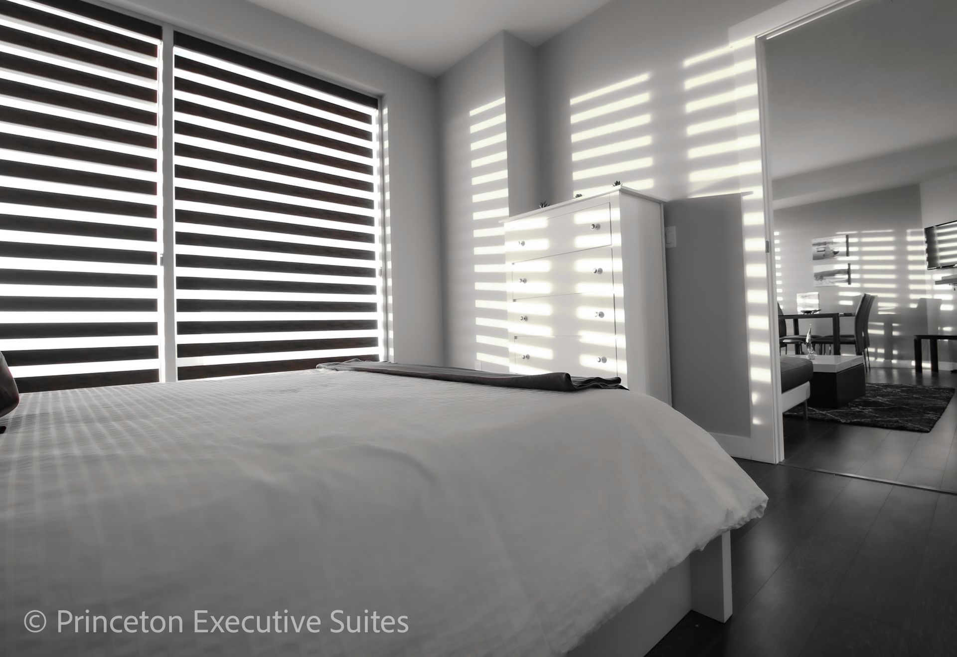 Bedroom with large windows, blinds, bed, and dresser. Neutral tones and natural light.